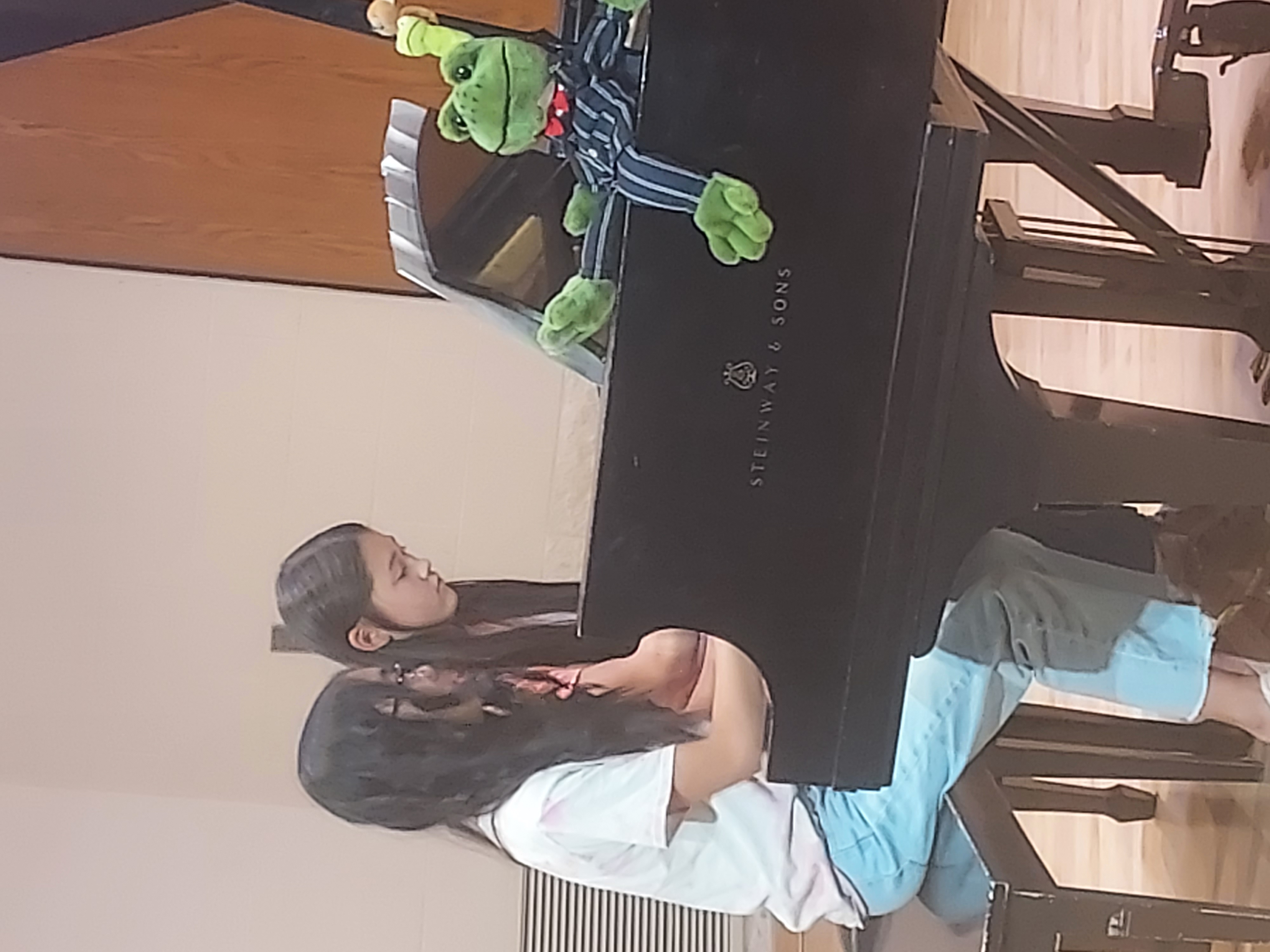 Picture of two girls playing the piano next to a stuffed frog